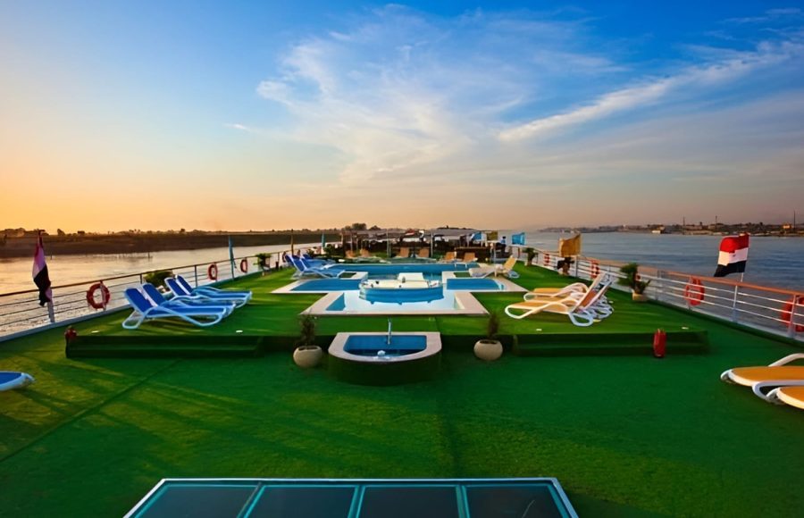 Wide view of a cruise ship sun deck during the day with a pool, green turf, and lounge chairs overlooking the Nile River.