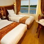 A twin-bed cabin on a cruise ship featuring white linens, bronze accents, and a large window with a view of the Nile River.