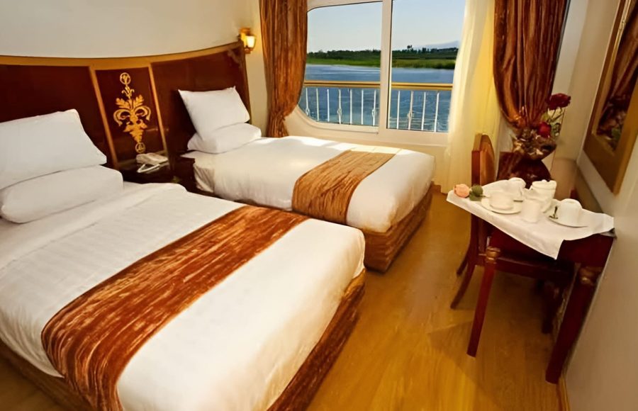 A twin-bed cabin on a cruise ship featuring white linens, bronze accents, and a large window with a view of the Nile River.
