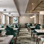 Modern cruise ship restaurant with marble tables, teal velvet chairs, and a buffet area.