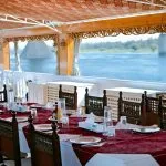 A long dining table with red and white linens set on a shaded wooden boat deck.