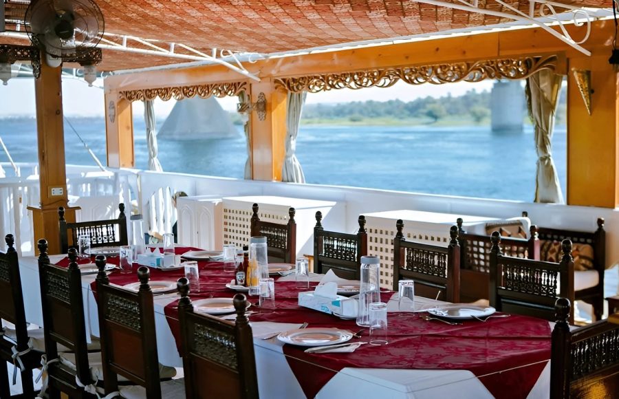 A long dining table with red and white linens set on a shaded wooden boat deck.