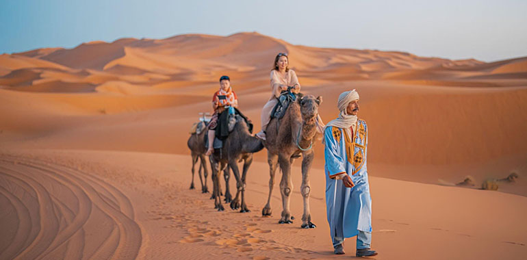 Asian Chinese Female Tourist Camel caravan going through the Sahara desert in Morocco at sunset lead by camel driver