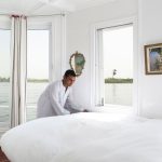 A cabin crew member in a white galabeya making a bed inside a bright, white Nile cruise cabin with large windows overlooking the river.