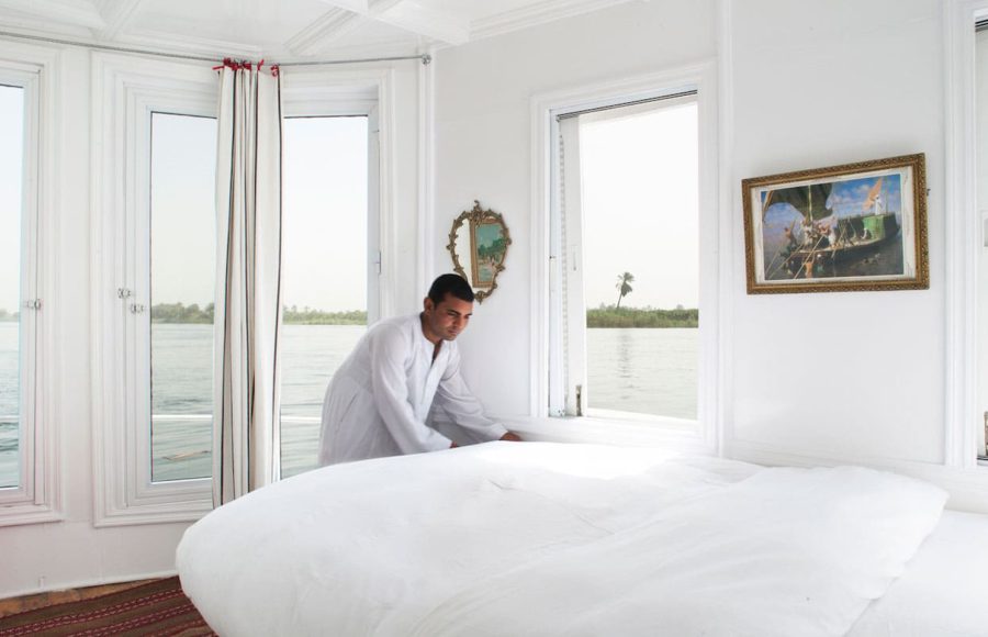 A cabin crew member in a white galabeya making a bed inside a bright, white Nile cruise cabin with large windows overlooking the river.