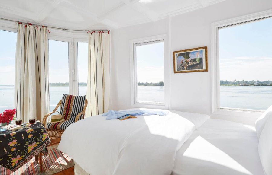 A bright, white cabin interior of a Dahabiya Nile cruise boat featuring a bed with a river view.