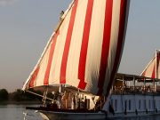 A traditional Egyptian Dahabiya boat with large red and white striped sails sailing on the Nile at sunset.