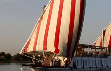 A traditional Egyptian Dahabiya boat with large red and white striped sails sailing on the Nile at sunset.
