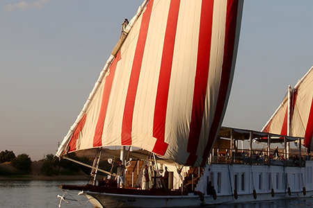 A traditional Egyptian Dahabiya boat with large red and white striped sails sailing on the Nile at sunset.