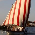 A traditional Egyptian Dahabiya boat with large red and white striped sails sailing on the Nile at sunset.