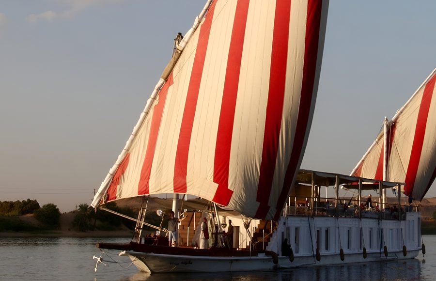 A traditional Egyptian Dahabiya boat with large red and white striped sails sailing on the Nile at sunset.