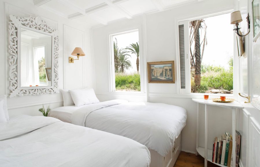 Two twin beds in a bright, white cabin on a Nile riverboat with large windows overlooking palm trees.