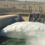 A large volume of white, foamy water being released from the lower sluices of the Aswan High Dam structure into the Nile River below.