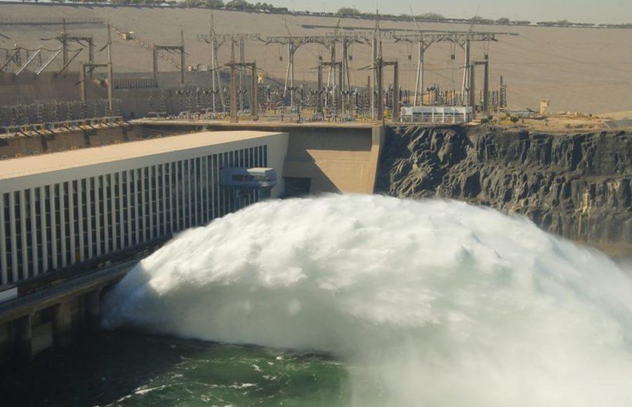 A large volume of white, foamy water being released from the lower sluices of the Aswan High Dam structure into the Nile River below.