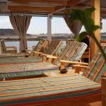 A row of wooden lounge chairs with colorful striped cushions on a shaded boat deck overlooking the Nile.
