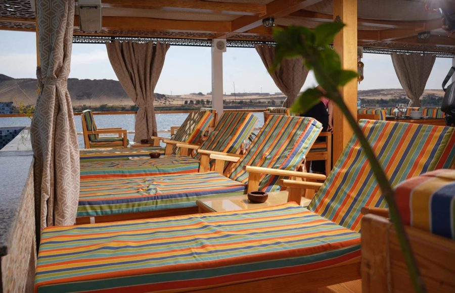 A row of wooden lounge chairs with colorful striped cushions on a shaded boat deck overlooking the Nile.