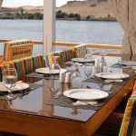 A glass-topped dining table set with white plates and glassware on a cruise deck.