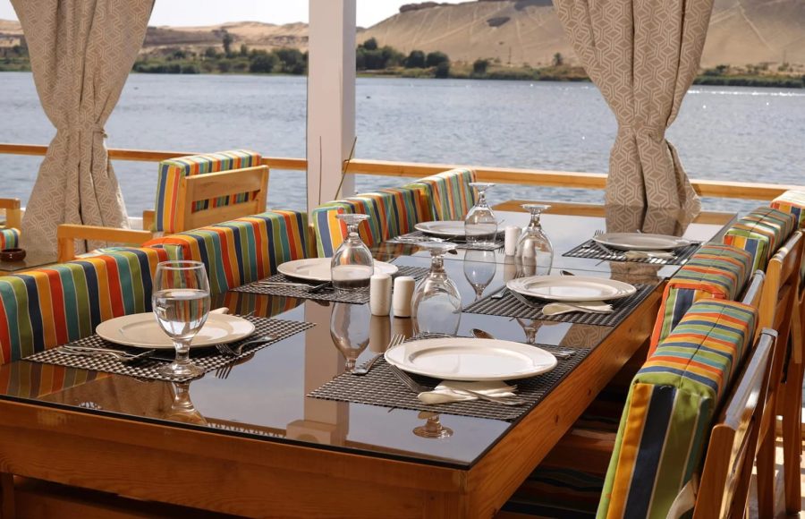 A glass-topped dining table set with white plates and glassware on a cruise deck.