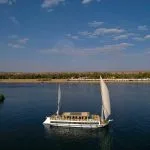 An aerial shot of a traditional white Dahabiya sailing vessel on the blue water of the Nile.