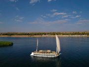 An aerial shot of a traditional white Dahabiya sailing vessel on the blue water of the Nile.