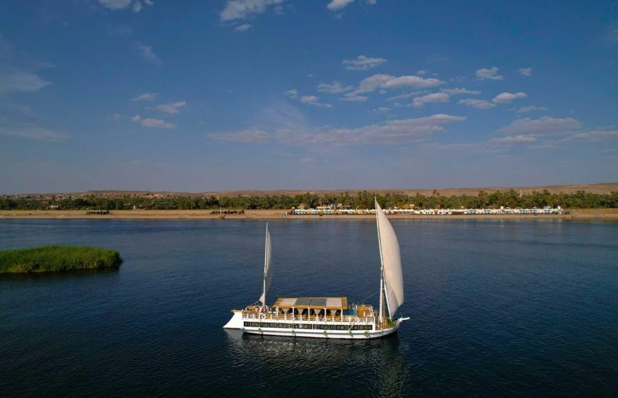 An aerial shot of a traditional white Dahabiya sailing vessel on the blue water of the Nile.