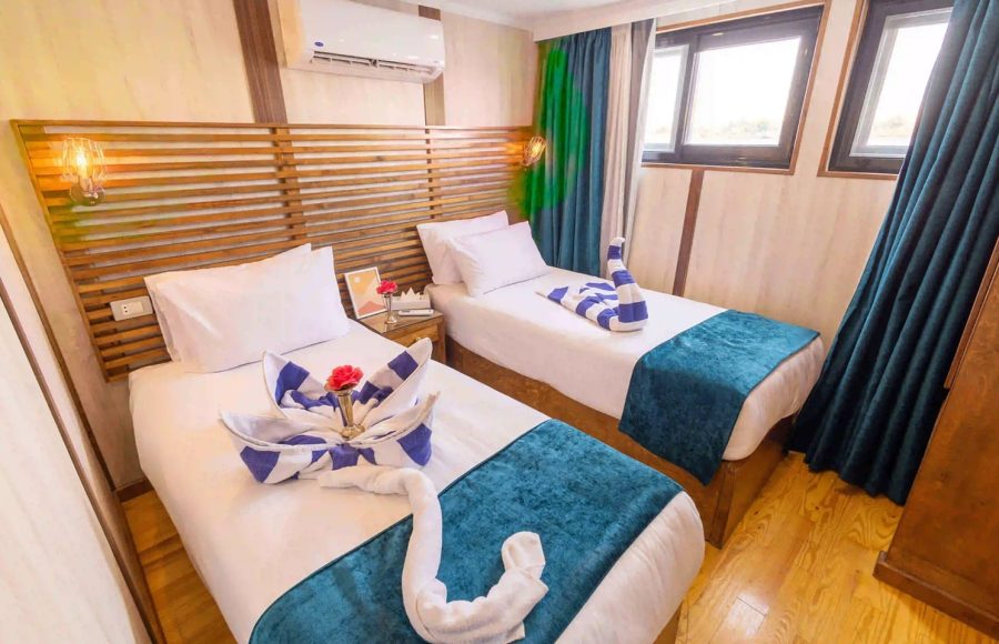 A cruise ship cabin with two twin beds featuring decorative towel art and blue velvet runners.