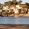 A cluster of colorful stone and brick houses on the sunlit bank of the Nile River in Egypt, framed by palm trees.