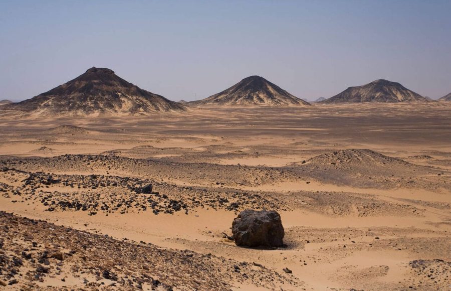 A wide panoramic view of the Black Desert Located in Bahariya Oasis in Western desert of Egypt, showing several distinctive, dark, conical, volcanic hills rising from the sandy, rocky ground.