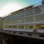 A close-up, low-angle view of the white, multi-deck M.S. Nile Palace cruise ship, showcasing its rows of large, gold-tinted windows reflecting the water and sunlight.