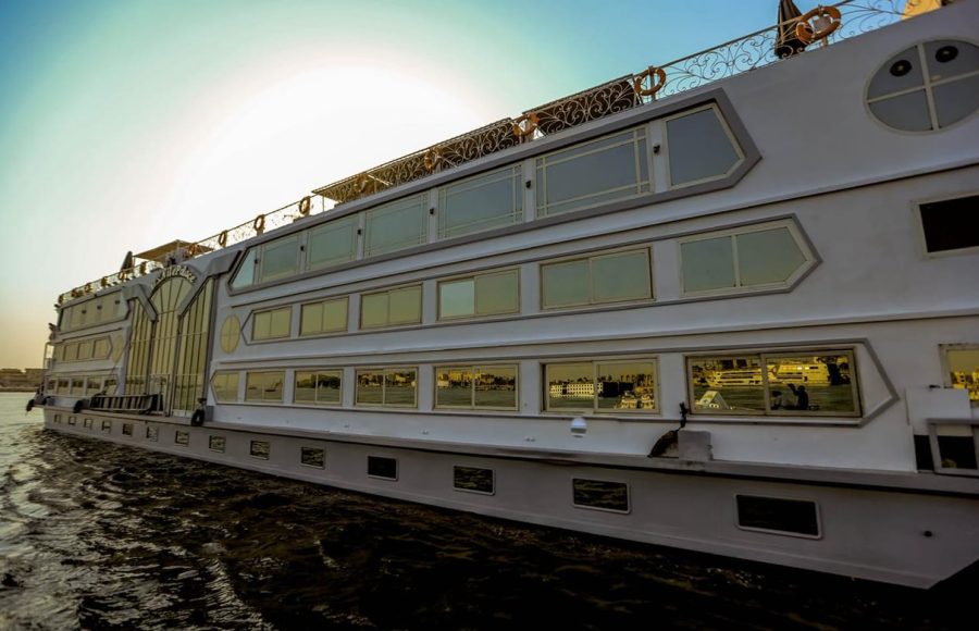 A close-up, low-angle view of the white, multi-deck M.S. Nile Palace cruise ship, showcasing its rows of large, gold-tinted windows reflecting the water and sunlight.