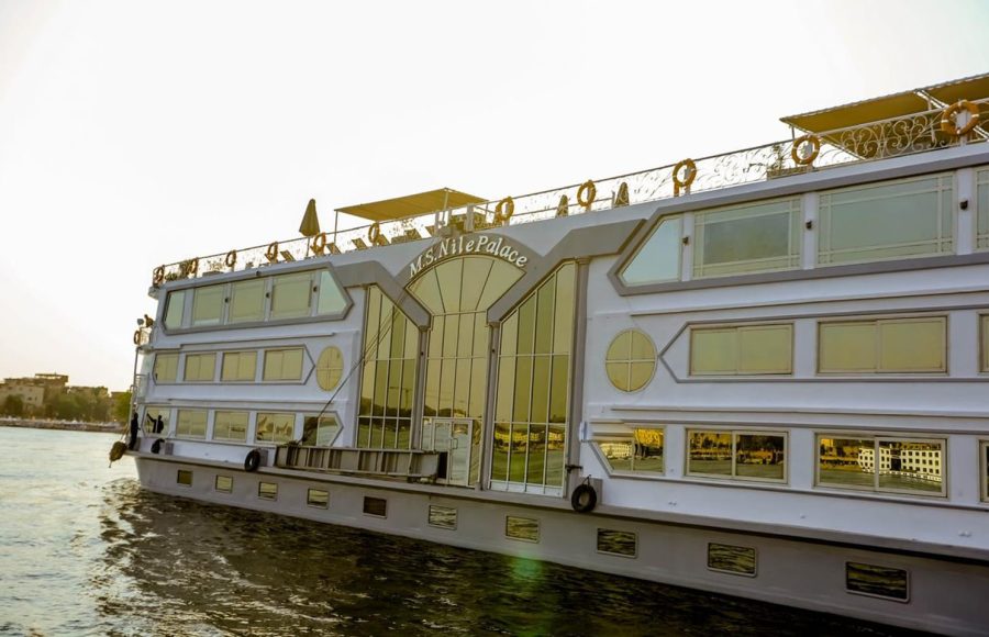 The port side of the white, multi-deck M.S. Nile Palace cruise ship, featuring large, gold-tinted windows, sailing on the Nile River at sunset.