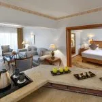 The interior of a spacious suite on the Nile Palace Deluxe cruise ship, showing a living room and a separate bedroom. The living area has grey sofas, a glass coffee table, and large windows with sheer white curtains. In the foreground, there is a counter with snacks, a kettle, and cups. The bedroom, visible through a wooden doorway, contains a large bed with a light wood headboard.