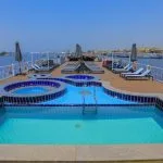 The main swimming pool and surrounding deck of the Nile Palace Deluxe cruise ship. The pool is a unique shape, lined with blue and white tiles, with two adjacent circular hot tubs. The wooden deck is surrounded by a white railing and features rows of blue lounge chairs and sun umbrellas. The ship is sailing on the Nile River, with a cityscape visible in the background.