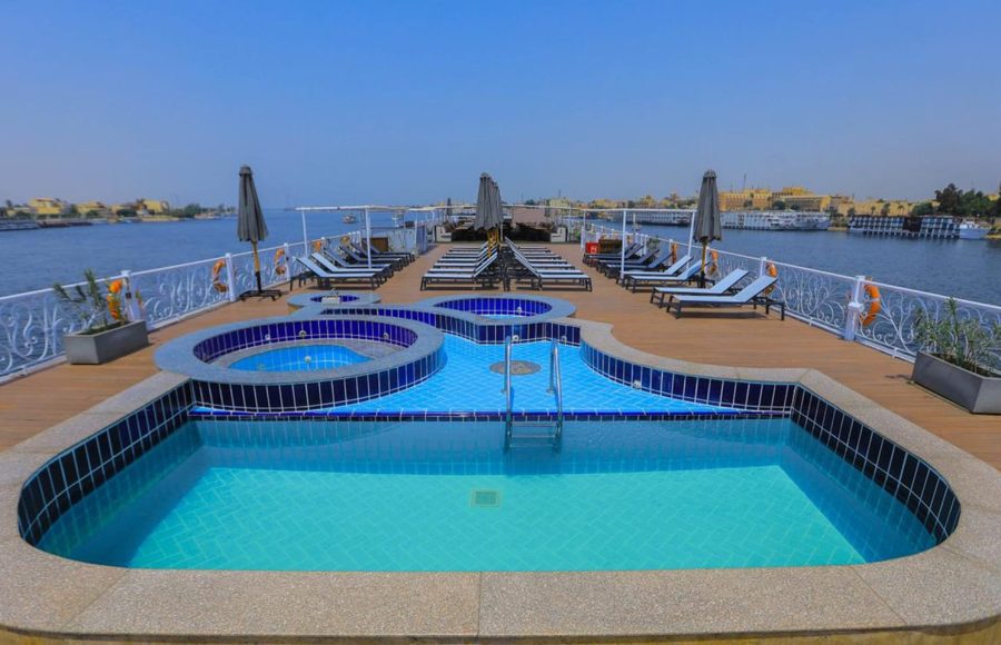The main swimming pool and surrounding deck of the Nile Palace Deluxe cruise ship. The pool is a unique shape, lined with blue and white tiles, with two adjacent circular hot tubs. The wooden deck is surrounded by a white railing and features rows of blue lounge chairs and sun umbrellas. The ship is sailing on the Nile River, with a cityscape visible in the background.