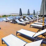 The sun deck of the Nile Palace Deluxe cruise ship, featuring rows of white and grey cushioned lounge chairs, black sun umbrellas, and a wooden deck floor under a clear blue sky. A small hot tub is visible on the left, and the background shows the shore and hills along the Nile River.