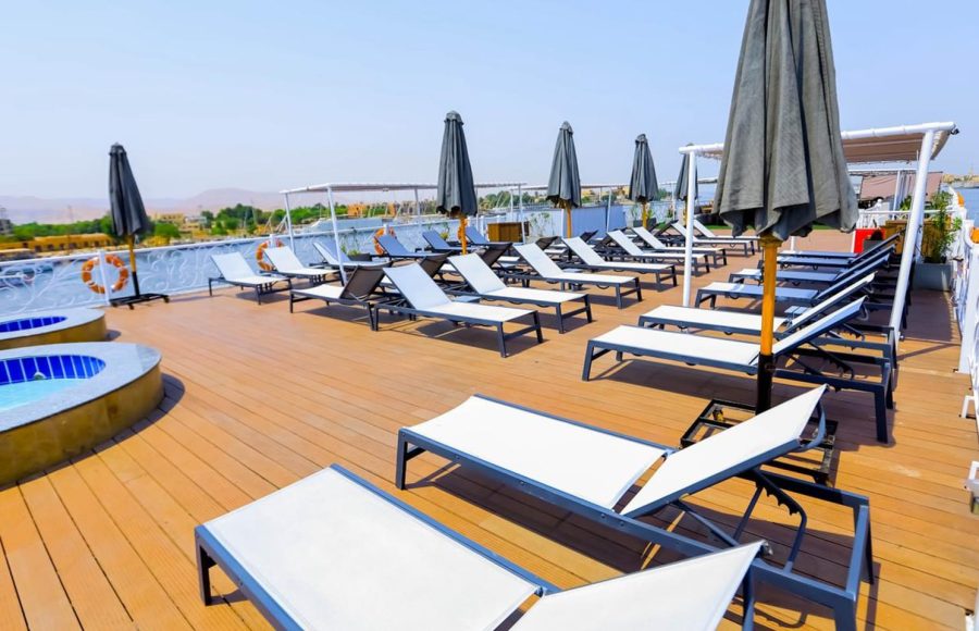 The sun deck of the Nile Palace Deluxe cruise ship, featuring rows of white and grey cushioned lounge chairs, black sun umbrellas, and a wooden deck floor under a clear blue sky. A small hot tub is visible on the left, and the background shows the shore and hills along the Nile River.