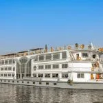 A wide exterior shot of the white, multi-deck "Nile Palace" cruise ship, featuring large rows of windows, sailing on the Nile River under a clear blue sky.