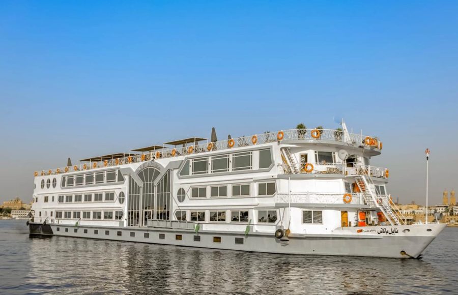 A wide exterior shot of the white, multi-deck "Nile Palace" cruise ship, featuring large rows of windows, sailing on the Nile River under a clear blue sky.