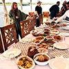 A long dining table on a boat deck filled with various Egyptian dishes, with people standing nearby and the Nile River in the background.