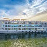 A wide exterior shot of the Cleopatra Nile Cruise ship, a multi-deck, white vessel with rows of blue-trimmed windows, sailing on the green waters of the Nile River under a dramatic sky.