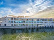 A wide exterior shot of the Cleopatra Nile Cruise ship, a multi-deck, white vessel with rows of blue-trimmed windows, sailing on the green waters of the Nile River under a dramatic sky.