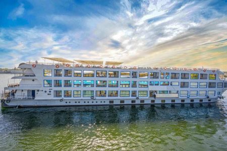 A Wide Exterior Shot Of The Cleopatra Nile Cruise Ship, A Multi-Deck, White Vessel With Rows Of Blue-Trimmed Windows, Sailing On The Green Waters Of The Nile River Under A Dramatic Sky.