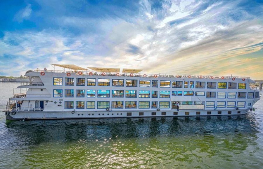 A wide exterior shot of the Cleopatra Nile Cruise ship, a multi-deck, white vessel with rows of blue-trimmed windows, sailing on the green waters of the Nile River under a dramatic sky.