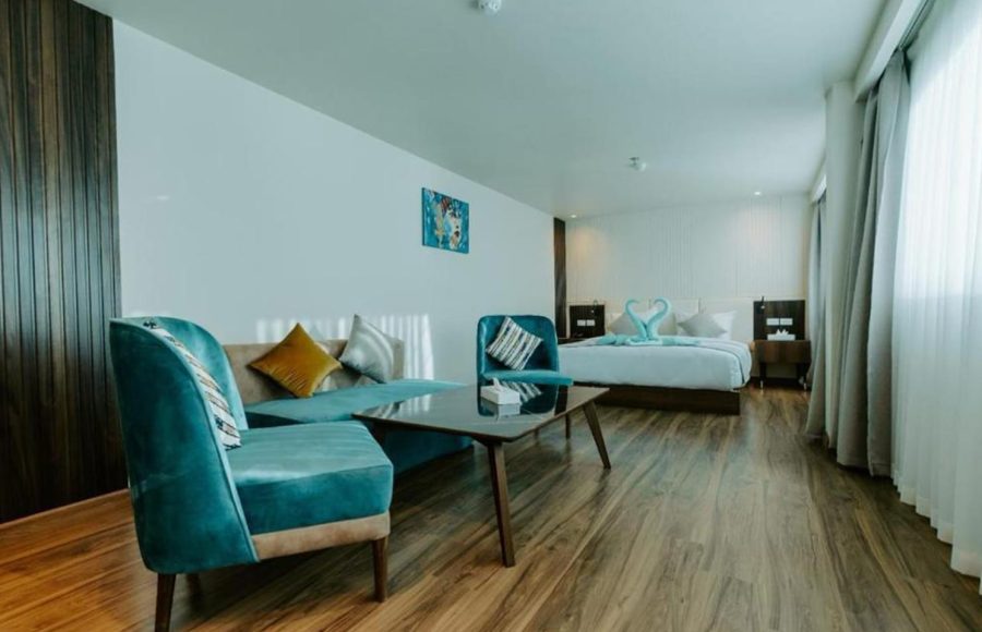 A spacious, modern Nile Cruise suite featuring a king-size bed in the background and a separate seating area with a sofa, two teal velvet armchairs, and a coffee table in the foreground.