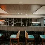 The main bar area on a Nile Cruise ship, featuring a wooden counter, a backlit shelving unit stocked with bottles, and four modern bar stools with beige backs and teal cushions.