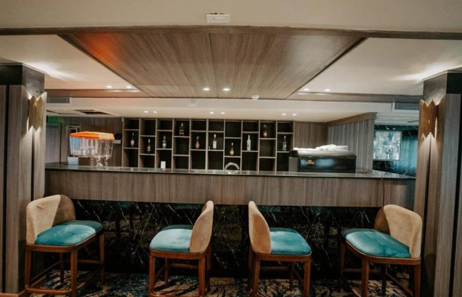 The main bar area on a Nile Cruise ship, featuring a wooden counter, a backlit shelving unit stocked with bottles, and four modern bar stools with beige backs and teal cushions.