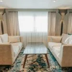 A contemporary cruise ship lounge area with two facing beige sofas, a geometric marble-top coffee table, and a colorful abstract carpet in blue and red tones.