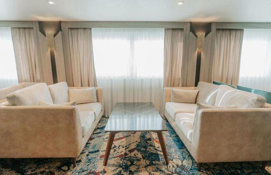 A contemporary cruise ship lounge area with two facing beige sofas, a geometric marble-top coffee table, and a colorful abstract carpet in blue and red tones.