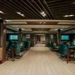 A wide-angle view of a modern, stylish Nile Cruise lounge with a light wooden central walkway, dark slatted ceiling, and various seating areas featuring beige sofas and deep teal club chairs.