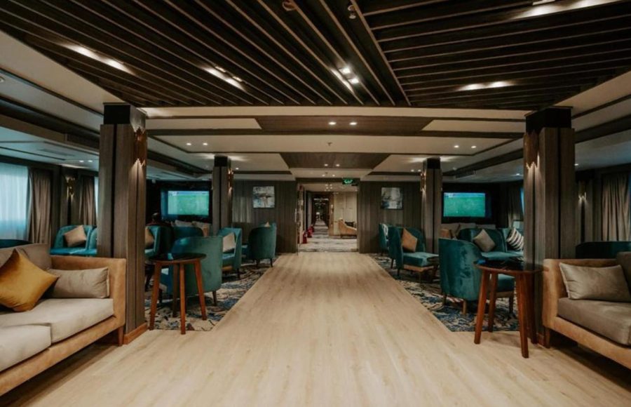 A wide-angle view of a modern, stylish Nile Cruise lounge with a light wooden central walkway, dark slatted ceiling, and various seating areas featuring beige sofas and deep teal club chairs.